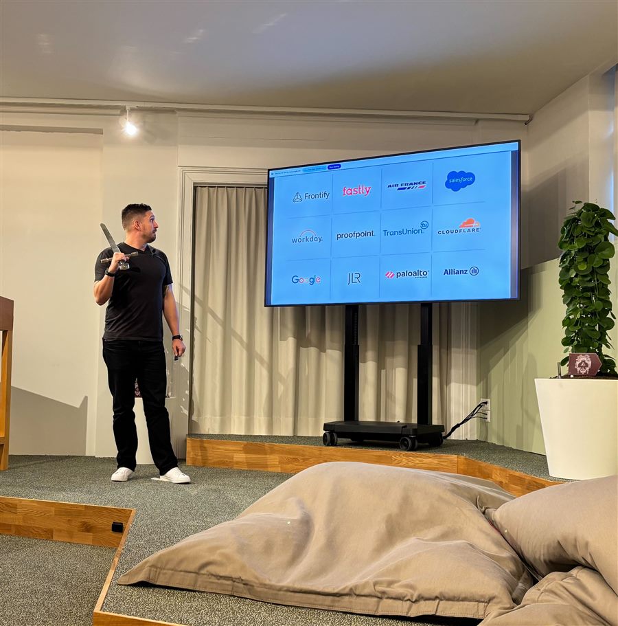 Peter giving a talk in front of a screen of customer logos, holding a sword over his shoulder.