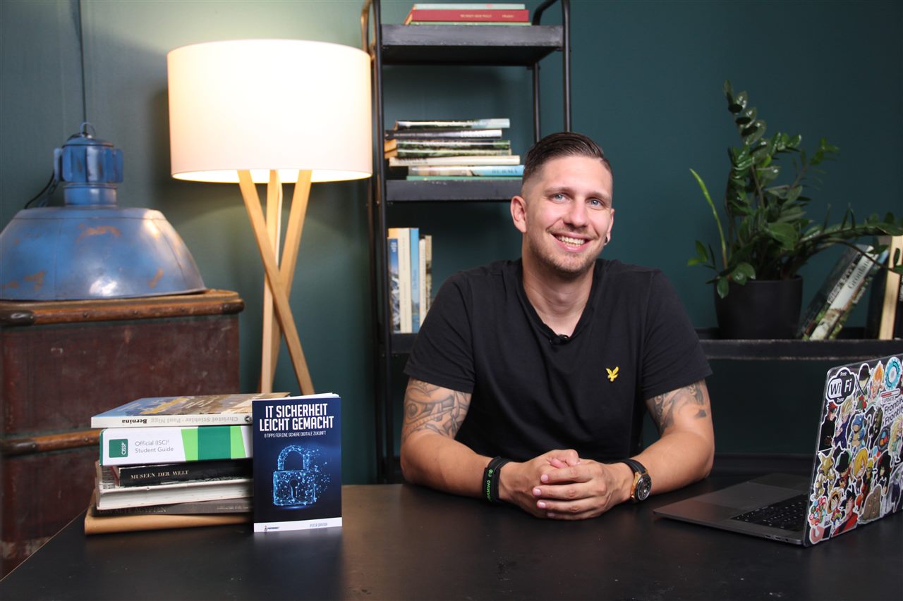 Peter Davida sitting at a desk in a green room, smiling, with a stack of books, a laptop and a copy of his e-book beside him.