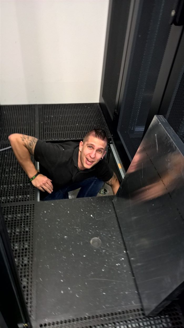 Peter grinning up at the camera from under a raised datacenter floor, next to a server rack.