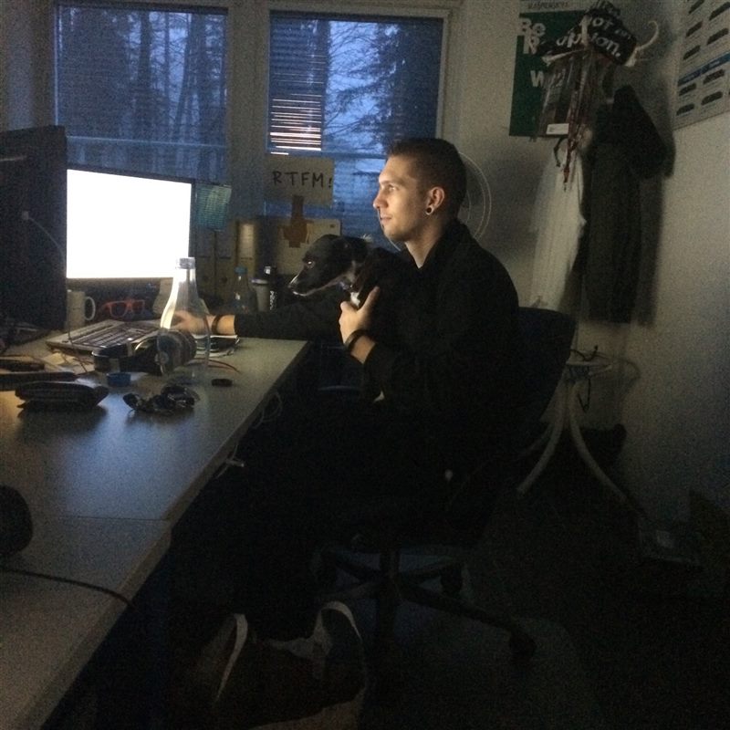 Peter at a dark office desk late in the evening, holding a small black dog, with a sign reading “RTFM!” on the wall.