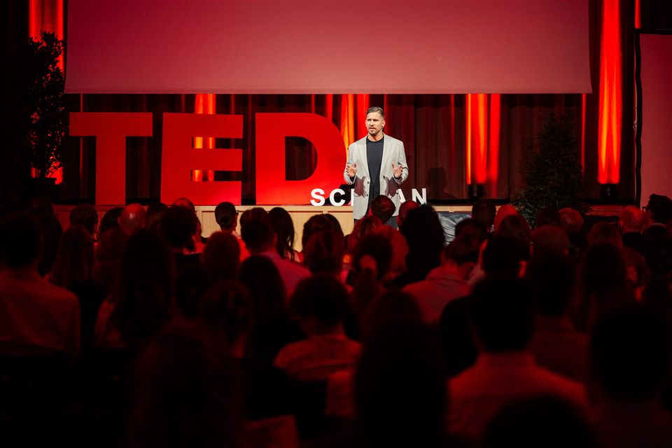 Peter on the TEDxSchaan stage in front of large red TED letters, speaking to a full audience.